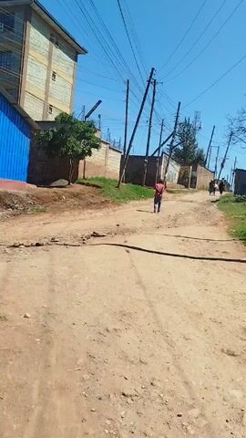 The video shows a dirt road in a developing area. A person in a red shirt walks away from the camera on the road. Other people are visible further up the road, walking in the same direction. The road is cracked and uneven. On the left side of the road is a building with a blue exterior and a larger building with multiple stories. There's a short wall lining the road, with grass growing between it and the road. Utility poles with wires run along the side of the road. On the right side of the road, there's a grassy area and a few buildings with corrugated iron roofs. The sky is clear and blue. The overall impression is of a low-income residential area.
