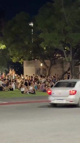 At night, a large group of people is seen sitting on the grass in a park-like setting.  The park is bordered by a street. Several people sit on low cement walls bordering the grass. A bicycle is parked nearby. Streetlights illuminate the area. Tall buildings are visible in the background. A traffic light cycles from green to red. A few cars pass by.