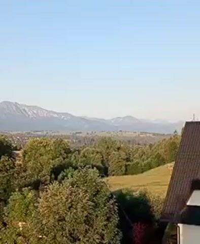 The video displays a vast landscape from a high vantage point. In the foreground, the dark roof of a building is partially visible on the right, suggesting the viewer is on a balcony or upper floor. A dense cluster of green trees dominates the middle ground, their leaves illuminated by the sun. Beyond the trees, a rolling green hill slopes upwards. In the distance, a sprawling town or village is nestled in a valley, with buildings visible amongst more trees. The backdrop is a majestic mountain range, with snow-capped peaks under a clear, pale blue sky. The lighting suggests it is either early morning or late afternoon. There are no people or animals visible in the frame. The camera angle appears to be slightly tilted upwards.
