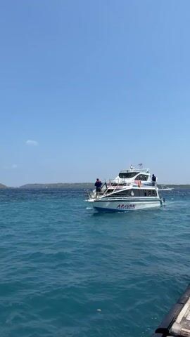 A modern white and blue passenger boat named "ARJUNA" travels across clear blue water under a bright blue sky. The boat has multiple decks and windows, and appears to be moving at a moderate speed. Two people are visible on the deck of the boat. One person, wearing a blue shirt and dark pants, is standing near the front of the boat, looking out towards the water. Another person, wearing a blue shirt, is visible further back on the boat. The water is a vibrant turquoise, with small waves indicating a gentle breeze. In the distance, a hilly, green landmass is visible, suggesting the boat is traveling in a coastal area or between islands. The overall impression is one of a pleasant, sunny day on the water.