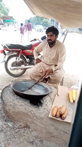 A man is squatting and stirring a large wok filled with what appears to be sand, on a small open-air stove made of mud. Next to the wok is a piece of cardboard with four ears of corn on it. Behind the man is a red motorcycle and a street with people and cars passing by. The man is wearing a traditional tan outfit and has a beard. He looks up at the camera and smiles. He appears to be preparing to roast the corn.