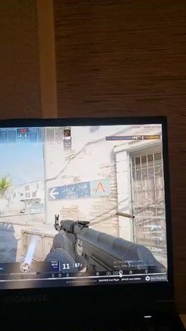 The video shows a laptop screen displaying a first-person shooter game, likely Counter-Strike. The player is holding an AK-47 and firing at enemies in a Middle Eastern-themed urban environment. A sign for "Hotel Aurore" is visible on the wall. The player's health and ammunition are displayed in the corner. The laptop is a Gigabyte brand. The room is dimly lit.  The person holding the camera is static.