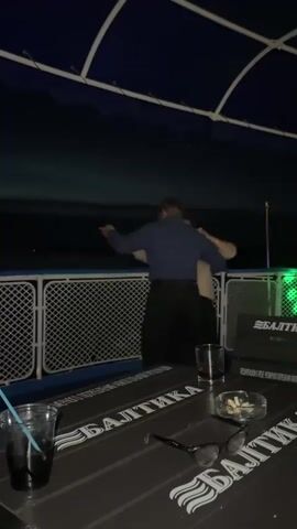 A couple is embracing and dancing on a boat deck at night. The man, wearing a blue shirt and black pants, has his arms around the woman, who is wearing a beige sweater and black pants. They are facing away from the camera. In the foreground, a table is visible with two drinks, a pair of glasses, and an ashtray with cigarette butts. The table has text that reads "БАЛТИКА". The background shows a dark body of water and a fence along the edge of the deck. The overall lighting is dim, with some colored lights visible on the boat.