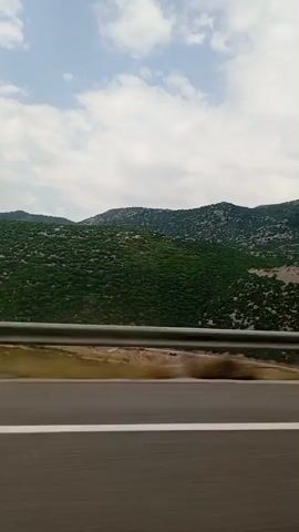 The video shows a side view from a vehicle traveling on a road. In the foreground, there is a dark asphalt road with a white line marking its edge. A silver guardrail runs horizontally across the middle of the frame. Beyond the guardrail, there is a strip of yellowish-brown ground with sparse vegetation. In the background, a large, rolling hill covered in dense green foliage is visible. The hill is dotted with lighter patches, suggesting exposed rock or less dense vegetation in certain areas. The sky above is partly cloudy with a mix of white and gray clouds. The image appears to be captured during daylight. The motion blur indicates the vehicle is in motion.