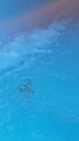 The video shows a close-up, downward-angled view of bright blue water. A person, mostly submerged, is visible in the water, appearing to be sliding down a water slide. The water is turbulent, with white foamy splashes and ripples, suggesting movement and speed. The environment appears to be a swimming pool or water park.