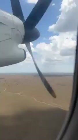 The video shows a view from the window of a small airplane. The spinning propeller is visible, and the landscape below is arid and brown, with sparse vegetation and a network of dirt roads. The sky is blue with scattered white clouds.