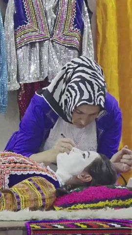 In this close-up shot, a woman with a black and white zebra-patterned hijab is applying makeup to a young girl. The woman, wearing a vibrant purple jacket over a white garment, leans over the girl, who is lying down with a white cream or mask on her face. The woman is carefully using a small brush to apply makeup to the girl's eyebrow. Both subjects are adorned in colorful, traditional Bulgarian attire. The girl wears a dress with intricate patterns in shades of yellow, orange, purple, and pink. The background features several other examples of richly decorated clothing, including jackets adorned with sequins and embroidery in blue and silver, and a bright yellow fabric hanging prominently. The scene appears to be an indoor setting, possibly a market stall or a dressing room, given the display of garments. The focus on the makeup application and the traditional clothing suggests an event or ritual, perhaps a preparation for a festival or celebration. The overall impression is one of care and tradition.