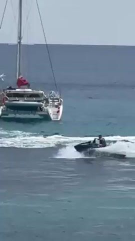 The video shows a black jet ski driven by a man near a white catamaran sailboat in the ocean. The ocean water is a mix of light and dark blues.  Two men are visible on the catamaran, one standing near the mast. The jet ski speeds through the water, creating a wake.  The catamaran is anchored and stationary. The sky is overcast.
