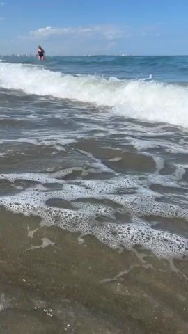 The video shows a close-up view of waves breaking on a sandy beach. The water is clear and the sand is a mix of pebbles and finer grains. In the background, a person is standing in the ocean, with the sky and clouds visible above. The sun appears to be shining, casting light on the water and the beach. The overall scene is one of a calm and peaceful day at the beach.