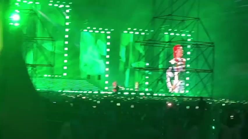 A concert is taking place in a large arena. Green lights illuminate the stage and the crowd. A large screen on the stage displays a person in a colorful outfit. The crowd is holding up phones with their flashlights on, creating a sea of lights. Some parts of the arena are dark, while others are brightly lit by the stage lights. The words "AK BARS ARENA" are visible on the upper sections of the arena.
