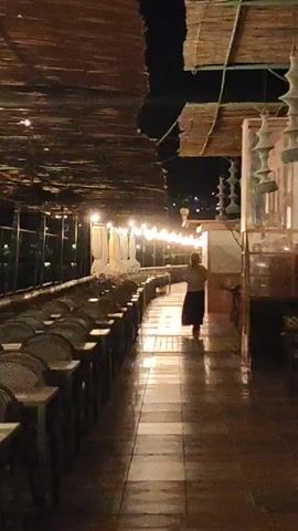 This night-time video captures a long, semi-outdoor dining or lounge area. The setting appears to be a restaurant or bar, possibly in Dénia, Spain, characterized by a rustic yet stylish ambiance. The overhead structure is a canopy or roof made of woven natural materials, casting intricate shadows.

On the left side of the frame, numerous empty chairs with a wicker or rattan design are neatly arranged along a long, low table, extending into the distance. The seating area is deserted, suggesting either very late or early hours, or a quiet moment.

The pathway in the center of the frame is a tiled floor, reflecting the warm glow of string lights strung overhead. These exposed bulb lights provide the primary illumination, creating a cozy and inviting atmosphere. In the distance, walking away from the camera, is a single person dressed in dark pants and a lighter-colored top. Their activity is simply walking through the space.

On the right side of the frame, there are structural elements, possibly glass partitions, and decorative hanging light fixtures or ornaments that complement the natural aesthetic. Beyond the immediate covered area, more artificial lights are visible in the background, hinting at a wider outdoor space or view. The overall impression is one of quietude and calm, with the warm lighting creating a peaceful environment. There is no visible text in the video.