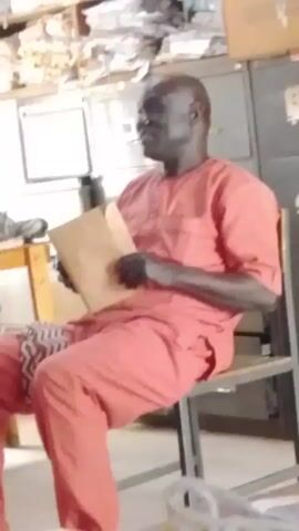 The video shows a man sitting on a chair and holding a document. He is wearing a red outfit. The background features a cluttered office space with cabinets, papers, and a table. He looks at the document and then looks to the side, smiling slightly.