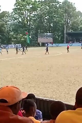 The video shows a rugby match in progress from the perspective of someone in the stands. Players in blue and red jerseys are scattered across a grass field. Trees surround the field, and there's a scoreboard and stands visible in the background. People are seated in the stands, and a person wearing a black hoodie with a graphic design on the back walks in front of the camera.