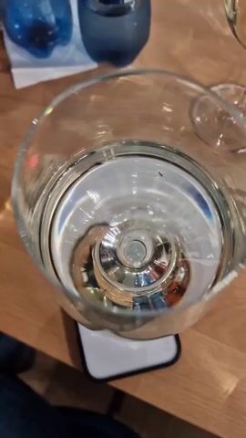 The video shows a wine glass filled with clear liquid sitting on top of a phone. The glass is large, with a wide bowl and a narrow stem. The liquid appears to be water or white wine. Bubbles form and dissolve in the water, and the light reflections off the glass create patterns. The phone underneath the glass is white and black with multiple camera lenses on its back. The table is made of light brown wood. In the background, there are two blue glasses. The video's angle is from above, looking down into the glass.