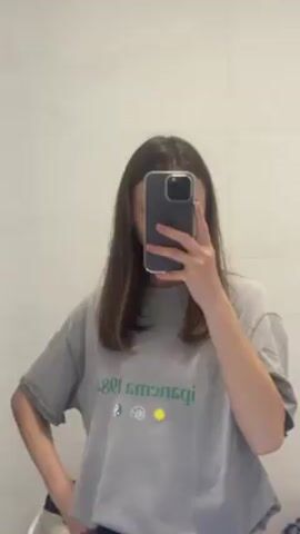 A woman with straight, shoulder-length brown hair stands in front of a bathroom mirror, taking a selfie with her smartphone. Her face is obscured by the phone. She is wearing a light gray t-shirt with a graphic design printed on the front in green, featuring text and symbols. She is also wearing dark bottoms. The bathroom has white walls and a toilet is partially visible in the lower-left corner.  The lighting is bright and even.