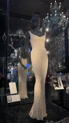 The video shows a display of Marilyn Monroe's iconic "Happy Birthday Mr. President" dress, encrusted with thousands of rhinestones, on a mannequin within a museum exhibit. The dress is a pale, form-fitting sheath with a low back. The exhibit features mirrored walls and a black, sparkling floor, creating a dazzling, reflective environment. In the background, partially obscured by reflections and glass, are glimpses of other museum displays, including framed photographs and possibly jewelry. A sign with a portrait of John F. Kennedy and text indicating "President Kennedy's Birthday Gala All Star Show" is visible near the dress. Several people are present in the background, some appearing to be museum visitors interacting with the exhibit, and one person, possibly a museum attendant, is seen in the distance. The lighting is bright and dramatic, emphasizing the sparkle of the dress and the surrounding decor.