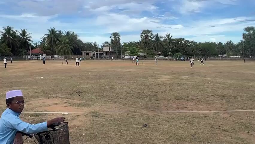 A wide outdoor shot depicts a cricket match taking place on a dry, grassy field. In the foreground, a young boy wearing glasses and a white prayer cap sits on a bicycle, observing the game. The cricket field is occupied by multiple players in cricket whites, engaging in the game. Tall palm trees line the perimeter of the field, interspersed with a few buildings. The sky above is a mix of blue and cloudy patches. The camera remains static throughout the video.
