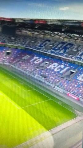This media file shows a wide, aerial view of a stadium's green grass football pitch. The pitch is neatly mowed with visible markings for the game. The stadium seating, primarily in blue and red, is largely empty. The upper sections of the stadium also show some seating in blue and red. Above the seating, there is a structure with various banners and signage.

The scene suggests a time of day when the sun is casting a strong shadow across the pitch, indicating it is likely daytime. The sky is visible at the top of the frame and appears to be clear blue. There are no people visible on the pitch or in the stands, suggesting the stadium is either empty or the event has not yet begun or has concluded.

Notable text visible in the media includes names and words displayed on the banners above the stadium seating. Some of these appear to be Polish names such as "SZARMACH," "PALASZ," "MATYSZ," and "DANKOWSKI," along with other words like "KRÓLEWSKIE" and "RZECZE." The overall impression is of a well-maintained, modern football stadium on a clear day, likely in Chorzów, Poland, given the location context.
