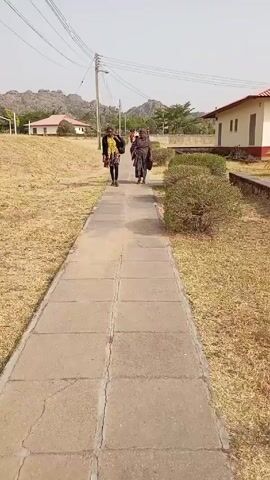Two women walk towards the camera on a concrete slab path, bordered by brown grass and small shrubs. The woman on the left wears a yellow print dress and black jacket, carrying a black bag over her shoulder. The woman on the right wears patterned clothing and carries a purse. Behind them are two buildings, distant mountains, and utility poles.