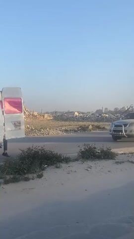 The video depicts a daylight scene on a paved road in a-Zawayda, Palestinian Territory. In the background, a vast mound of concrete and rebar rubble dominates the landscape, indicative of a destroyed large structure. The sky is clear and bright blue.

Two light-colored pickup trucks drive from left to right across the frame. The first, a modern-looking pickup with a prominent bull bar on its front, carries several men standing in its open bed, some holding onto the cab. Shortly after, a second, older model pickup truck passes. Its bed is heavily crowded with numerous men, some standing, some seated, and one appearing to climb onto the side. This second truck is also towing a small, white, enclosed trailer.

The overall environment appears arid, with sandy shoulders beside the road. Throughout the video, upbeat music plays, accompanying the movement of people against a backdrop of extensive destruction under a clear sky.