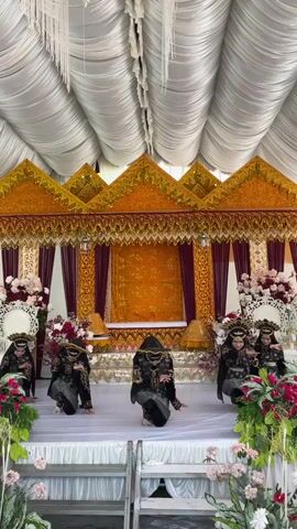 This is a vertical, eye-level shot of a group of five young women in traditional attire performing a dance on a stage. The stage is decorated with white fabric and flanked by large floral arrangements featuring pink and red flowers. Behind the dancers, there is an ornate, gold-trimmed backdrop with triangular peaks and maroon curtains. The ceiling above is draped with white fabric creating a tent-like effect. The dancers are all wearing matching black and silver outfits with intricate patterns, veils, and elaborate headpieces. They are positioned in a semi-circle, with one dancer in the center and two on each side. The central dancer is slightly more forward and appears to be leading the performance. The dancers' movements are graceful and synchronized, with some kneeling and others extending their arms. The overall atmosphere appears festive, possibly for a cultural event or celebration.