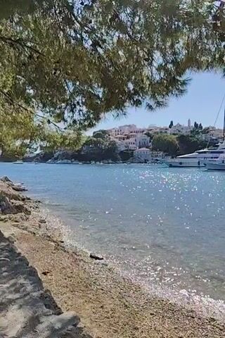 A video pans across a clear blue bay with several boats docked. In the foreground, a pebbled beach is visible with some rocks and a tree branch with green needles obscuring the top of the frame. The water is calm and transparent, allowing the seabed to be seen. Behind the boats, a white-washed town is nestled on a hillside, with buildings and trees dotting the landscape under a clear blue sky. The video captures a serene coastal scene with boats of various sizes, including a large white yacht. The overall impression is a picturesque Mediterranean harbor.