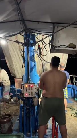 The video shows three men on a boat at night. Two men are visible in the background, and one man is in the foreground. The man in the foreground is shirtless and wearing shorts, facing away from the camera and standing next to a blue mechanical device. The man on the left is wearing a light-colored shirt and dark pants, and appears to be operating a winch. The man on the right is wearing a blue shirt and checkered shorts, and is standing behind the man in the foreground. The blue mechanical device has a control panel with several buttons and lights. A large fishing net is visible in the background. The overall impression is that the men are engaged in fishing-related activities on a boat at night.