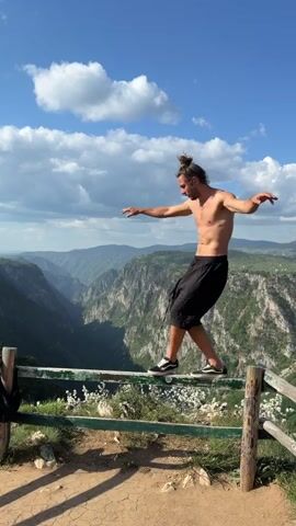A shirtless man with a bun is walking on a wooden fence in front of a mountainous landscape. He wears black shorts and black sneakers. The fence is old, with partially green and brown paint. White flowers are present near the bottom of the fence. He is balancing with his arms outstretched. The landscape consists of a deep canyon with trees and mountains in the distance. The sky is blue with scattered clouds.