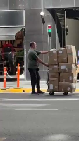 A man pushes a tall stack of brown cardboard boxes on a wheeled cart across a crosswalk in front of a store with a black awning. The store's sign reads "briganti." The man wears a green t-shirt and dark pants. The boxes have printed information on them. Traffic including cars, scooters and a bus drive by. Orange cones are along the curb. The crosswalk is marked with yellow paint. The traffic light is green. People walk in the background.