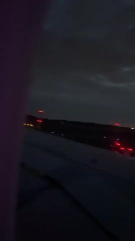 The video captures a nighttime airplane takeoff as viewed from a passenger window. The left wing of the aircraft occupies the lower portion of the frame, with a dark, indistinct object (possibly part of the window frame or an item held close to the camera) visible in the extreme left foreground, displaying a purple/pink hue.

The scene is an airport at night, under a heavily clouded, dark sky. Numerous bright white and yellow runway lights are visible on the ground, stretching into the distance. Several distinct red lights also punctuate the darkness. Beyond the airport, a faint glow of distant city lights can be seen on the horizon.

The primary activity is the airplane accelerating down the runway and taking flight. The video begins with the aircraft already in motion, rapidly passing the ground lights. The sound of powerful jet engines is audible, growing in intensity. Approximately halfway through the video, the plane smoothly lifts off the runway, and the ground lights gradually drop out of view as the aircraft ascends into the dark night sky. No people are visible, and no text appears in the frame.