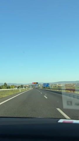 The video shows a view from inside a car driving on a highway under a clear blue sky. The road is asphalt with white lane markings. On the right side of the road, there are blue highway signs with symbols and text, including one indicating a distance of 750 meters and a route number. In the distance, there's an overhead gantry with a digital display sign. The landscape on either side of the highway appears to be rural with some trees and hills in the far distance. The interior of the car, specifically the dashboard area, is visible at the bottom of the frame. The video is shot from a fixed perspective, suggesting it's a recording of a journey.