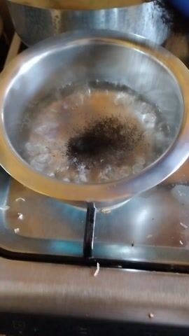 The video captures a top-down view of a metal pot on a gas stove, likely in a kitchen in Kikuyu, Kenya. Inside the pot, water is actively boiling, with dark, granular particles, resembling tea leaves or ground coffee, bubbling at the surface. The pot is made of shiny silver-colored metal. The gas stove's surface, also metallic, shows some small dark specks around the pot. The bubbling water indicates that the stove is on, though the flame itself is not visible.