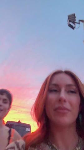 Two women record a selfie-style video in an outdoor setting with a vibrant sunset in the background. The woman on the right has long, auburn hair and wears a beige and brown patterned shirt, a layered necklace, and rings. She playfully sticks out her tongue and touches her forehead. The woman on the left has short, dark hair and wears a black top with orange trim and a silver pendant necklace. She smiles throughout the video. In the background, other people are milling around, along with what appears to be event infrastructure like stages, lights, and trucks.