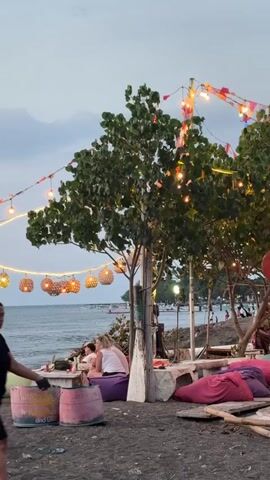 This media file depicts a beachside scene in Buleleng, Indonesia. The setting is a relaxed, open-air gathering space, likely a cafe or bar, situated on a dark sand beach. The time of day appears to be late afternoon or early evening, as indicated by the soft, diffused light and the illuminated string lights decorating the area.

Several people are present. In the foreground, a woman in a black top and shorts walks across the sand, her back to the camera. Further back, two individuals, possibly women, are seated on large purple beanbag chairs, engaged in conversation or relaxation. Another person is seated on the sand to the left, also in a relaxed posture. In the background, more people are visible along the shoreline, suggesting a public beach area.

The scene is characterized by a bohemian and casual aesthetic. Numerous string lights with woven globe shades are strung between trees and poles, casting a warm glow. Large beanbag chairs in various colors (purple, pink, green) are scattered around, along with low tables. Two repurposed industrial barrels, one pink and one teal with yellow accents, serve as decorative elements or perhaps impromptu seating. Trees with lush green foliage are interspersed throughout the seating area, adding to the natural ambiance. The ocean forms the backdrop, with gentle waves visible. The sky is overcast with a pale blue hue, suggesting cloudy weather.

The overall mood conveyed is one of leisure and tranquility. The soft lighting and comfortable seating arrangements invite relaxation. The presence of multiple people enjoying the space suggests a social gathering or a popular spot for unwinding. No discernible text is clearly visible in the media file.