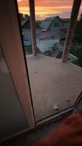 The video is a first-person perspective of someone looking out from a structure with two large wooden posts framing the view. The foreground is a concrete surface. Beyond the structure, a backyard is visible with green grass, trees, and several houses. The sky is filled with dramatic sunset clouds painted in shades of orange, pink, and yellow. There are no people or specific actions clearly identifiable in the short clip.