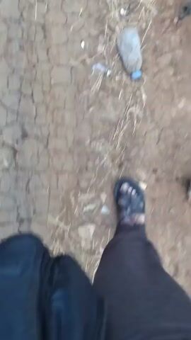 A first-person perspective video shows a person's legs and feet walking on a dry, cracked dirt path. The person is wearing dark pants and sandals. Scattered on the path are small pieces of debris, including what appears to be a plastic bottle. The camera is moving, suggesting the person is walking.
