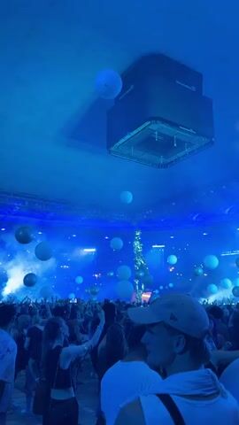 The video shows a crowded indoor event, likely a concert or festival, with a dark, blue-toned lighting scheme. A large, black speaker or lighting rig is suspended from the ceiling. Numerous white and gold balloons are floating throughout the venue, some appearing to be quite large. A tall, metallic sculpture or stage prop is visible in the distance, illuminated by spotlights. The crowd is densely packed, with many people holding up their phones, suggesting they are recording or taking pictures. The overall atmosphere appears to be energetic and festive.
