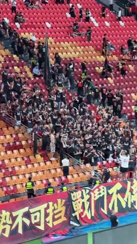The video shows a crowd of soccer fans at a stadium. The seats are red and orange. Most of the fans are wearing black shirts, with a few shirtless men in the foreground. They are standing in a block of seats, cheering, jumping, and waving their arms. A black flag is visible above the crowd. In front of the fans, there is a banner with Chinese characters on it. Security personnel in yellow vests stand in front of the banner, facing the crowd. Some people sit in the surrounding seats, watching the excitement below.