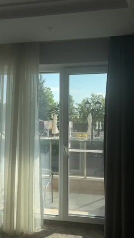 The video shows a view from inside a room, looking out through a glass door and window. The door is partially open, with sheer white curtains on the left and dark grey curtains on the right. Through the glass, a swimming pool area is visible. There are lounge chairs and white umbrellas surrounding the pool. Trees and a street lamp are also visible. The sky is clear and blue. The time stamp indicates the video starts at 00:00 and ends at 00:04.