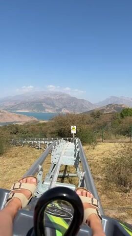 The video shows a first-person perspective of a person riding a roller coaster. The roller coaster is on a track that goes down a mountainside with a body of water and mountains in the background. The rider's feet, wearing sandals, are visible at the bottom of the frame. The sky is clear and blue.