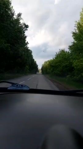 The video captures a first-person perspective from inside a vehicle driving along a two-lane paved road. The dashboard and windshield wipers are visible in the foreground, indicating the recording device is mounted within the car. The road stretches ahead, flanked on both sides by dense green trees, forming a natural canopy or tunnel effect. The sky above is largely overcast with bright clouds, suggesting daytime.

In the distance, a white car is seen approaching and then passing the recording vehicle, traveling in the opposite direction. The road appears to have some uneven patches or repairs visible. No other vehicles or people are distinctly visible. The overall setting is a rural or semi-rural area, consistent with the provided location context of Ablyazovo, Russia. There is no visible text. The scene conveys movement through a natural, green landscape under a cloudy sky.