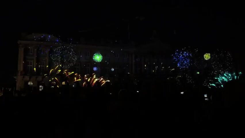 The video shows a nighttime fireworks display projected onto the facade of a large building. A crowd of people is visible in silhouette in the foreground, holding up glow sticks and phones. The fireworks are colorful and varied, with bursts of green, yellow, blue, purple, and red. The building facade itself is lit up with the projections, with some areas of the architecture also visible. The display appears to be a celebratory event, possibly a festival or holiday.