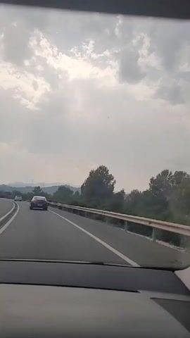 The video shows a view from inside a car as it travels on a highway. The road has multiple lanes and a metal guardrail runs along the right side. Outside the car, there are trees, vegetation, and rolling hills in the distance. The sky is cloudy, with patches of sunlight breaking through. There are other cars on the road, and in the distance, some wind turbines are visible. The scene appears to be a rural or semi-rural landscape.