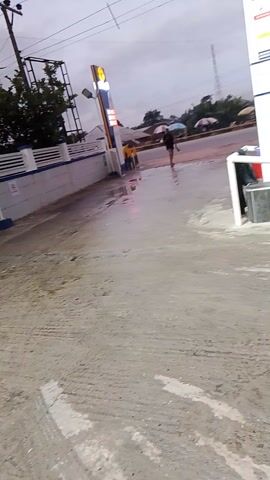 The video shows a gas station with two pumps visible. Pump number 1 is on the left and pump number 2 is to the right of it. A blue vehicle with the text "M1235" on its side is parked at pump number 2. A person wearing a red shirt and blue pants is standing at pump number 2, seemingly operating the pump. Another person wearing a brown shirt is standing behind them, also at pump number 2. The ground appears wet, suggesting it may have rained recently. The overall lighting suggests it is either dusk or a cloudy day.
