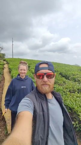 A man and woman are walking on a path through a tea plantation. The man is holding the camera and filming himself and the woman. The man is wearing a hat and sunglasses and has a beard. The woman is wearing a blue sweatshirt. They are both smiling. The tea plants are green and lush. The sky is cloudy. The woman puts her arm around the man.