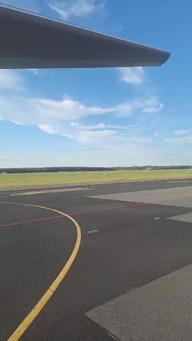 The video shows an airplane's wing and a runway. The camera appears to be inside the airplane, looking out the window. The runway has yellow and red markings. In the distance, another airplane is visible on the runway. The sky is blue with scattered white clouds. The landscape beyond the runway is green with trees in the distance. The video lasts for five seconds.