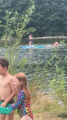 A person stands on a paddleboard in a lake, holding a paddle. Two other people are in the water nearby. A third person is in the water and appears to be jumping. Trees and foliage surround the lake.