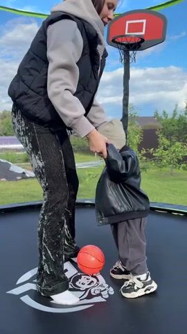 In an outdoor setting, a woman and a toddler stand on a trampoline. The woman, likely in her late twenties or early thirties, has dark hair and is wearing a gray hoodie, a black vest, and dark, patterned pants, along with white sneakers. She holds the hands of the toddler, who is dressed in a black leather jacket, gray pants, a beanie, and sneakers that are black, white, and yellow. A basketball rests on the trampoline near the child. Behind them, there is a basketball hoop attached to the trampoline. The trampoline is enclosed by a blue and green safety net. The background shows a green lawn, some trees, and a house in the distance.