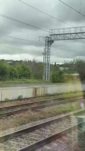 A view from a train window shows the train arriving at a station. The sky is overcast. The railway tracks are visible, with overhead power lines supported by metal structures. The station platform has a yellow safety line and a sign that reads "Perovskaya".