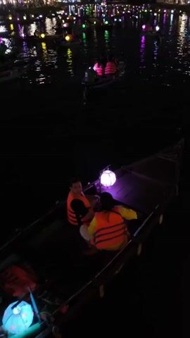 At night, a long narrow boat on a river is occupied by a man and a woman wearing orange life vests and two illuminated paper lanterns: one white and one turquoise. A rower in a green shirt boards the boat. In the background, other boats populate the water, with lanterns and lights reflecting on the water. Distant lights from buildings and trees line the riverbank.